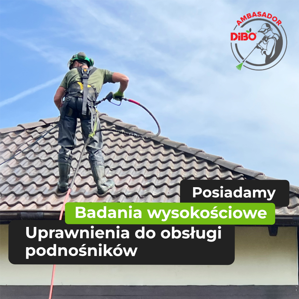 Pracownik w kasku i uprzęży bezpieczeństwa myje dach kryty dachówką cementową za pomocą myjki ciśnieniowej.